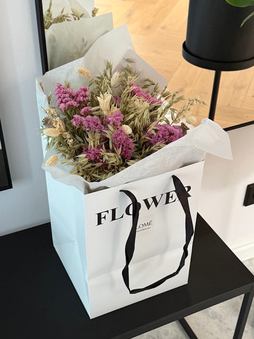 A white paper flower gift bag with black handles and the word 'FLOWER' printed on the front, filled with dried pink and beige flowers, sitting on a black table in a modern interior setting.