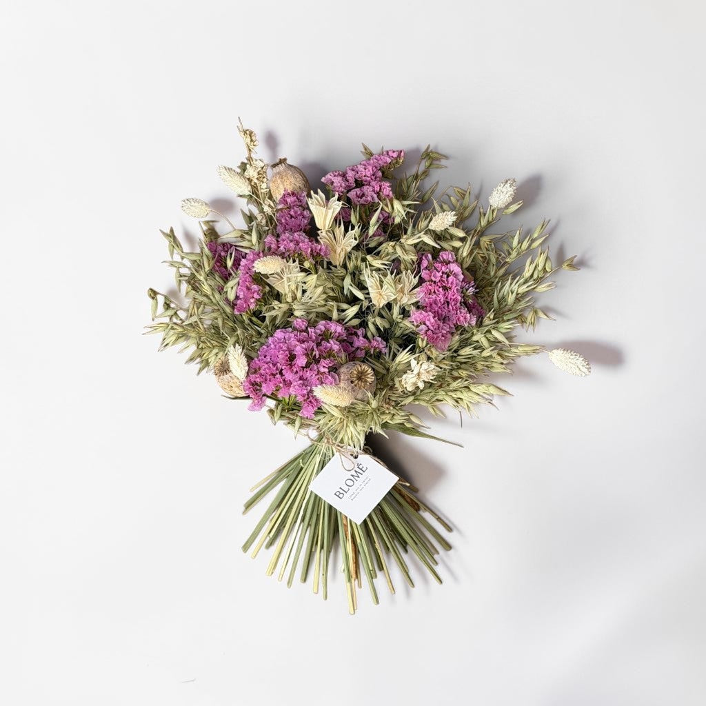 A dried flower bouquet with pink and purple blossoms, beige dried grasses, and green stems, tied with a white tag that reads 'BLOOM', photographed on a clean white background.